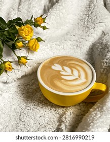 A cozy flat lay featuring a cup of latte with elegant latte art in a bright yellow ceramic cup, surrounded by fresh yellow roses on a soft white fluffy blanket. Warm, relaxing atmosphere perfect for themes of coffee, comfort, morning routine, self-care, relaxation, lifestyle, and hygge. Ideal with copy space for text.
