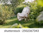 Cozy garden corner with wicker rocking chair. Calm outdoor seating for relaxation. Garden furniture against the background of lush plants. Garden planning and plant care. Horizontal photo