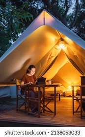 Cozy glamping tent with light inside and a woman using a laptop during dusk. Luxury camping tent for outdoor summer holiday and vacation. Lifestyle concept - Shutterstock ID 2138053033