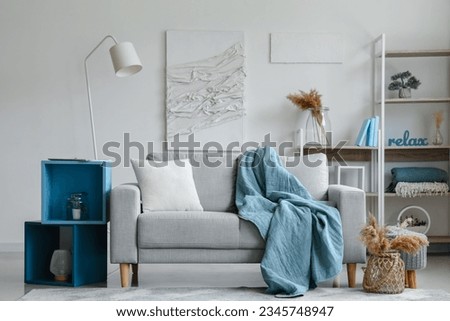 Cozy grey sofa with soft blanket, cushion and stylish shelves in interior of light living room