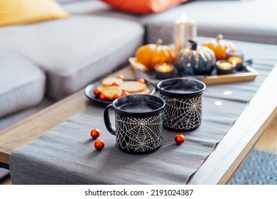 Cozy halloween plans at home. Hot tea drink in black mugs with spider net pattern, cookies and and sweets on the plate, pumpkin decor on the coffee table. Festive autumn, fall mood home decor