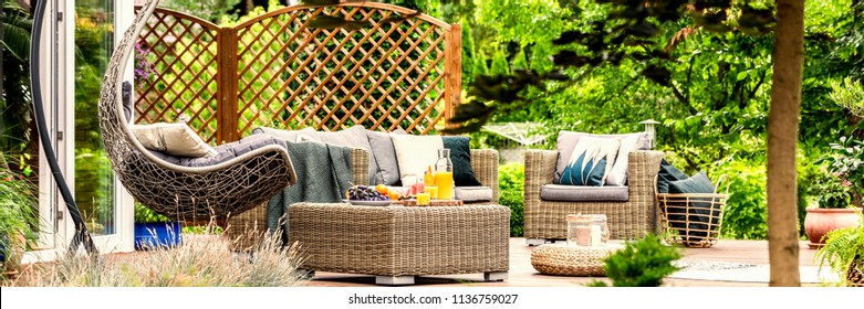 Cozy hanging egg chair, stylish garden furniture and fruit on a wicker table on a spacious terrace among green plants and trees