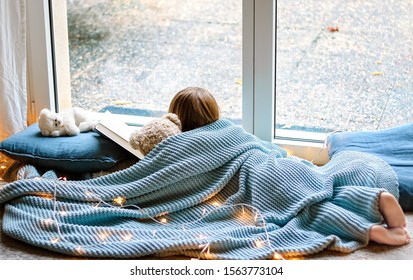 Cozy Holidays At Home. Cute Little Child Lying Under Blue Knitted Blanket With Teddy Bear Friend On Floor At Window Reading Book. Winter Season Lifestyle. Leisure Time.