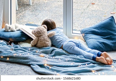 Cozy Holidays At Home. Cute Little Child Lying On Blue Knitted Blanket With Teddy Bear Friend Hugging Him On Floor At Window Reading Book. Winter Season Lifestyle. Leisure Time.