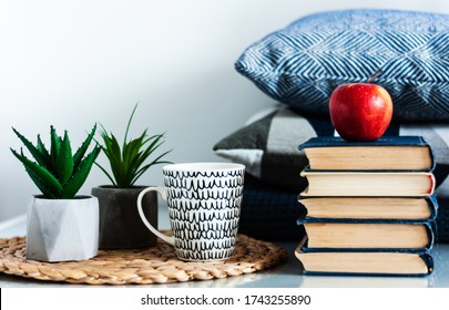 Cozy Home Interior Decor: Cup Of Coffee, Stack Of Books, Red Apple, Plants In Pots On A Wicker Stand, Blue Pillows On A White Table. Distance Home Education. Quarantine Concept Of Stay Home