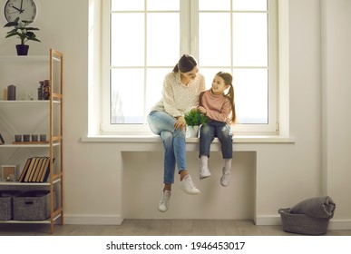 It's so cozy inside, let's stay at home. Happy family backlit by warm sunlight having fun in new house. Positive single mother together with little daughter sitting in sun on white plastic window sill