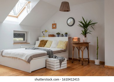Cozy interior in the attic. Bedrrom in apartment with clock on the white wall, double bed with stylish pillows, and wooden floor. Scandinavian design. Bright room hotel with simple decor.