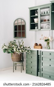 Cozy kitchen corner with vintage furniture of green colour, countertop with drawers, flowers in glass jar, hanging cabinet with glass doors, window shaped mirror, basket with greenery