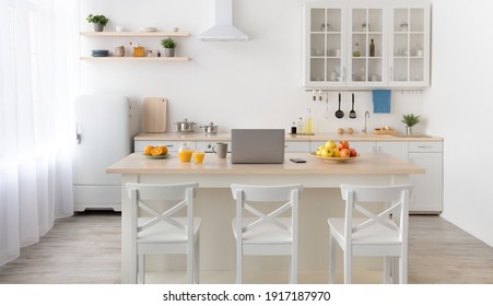 Cozy kitchen interior in morning, contemporary design. Plate with apples, cup, laptop and phone, glass and jug of orange juice on table, white chairs, utensils on furniture, light walls, curtains - Powered by Shutterstock