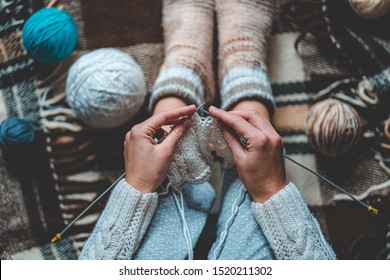 Cozy knitting woman in knitted winter warm socks and in pajamas enjoys knit work on brown checkered plaid blanket at home in cozy winter time. Top view 