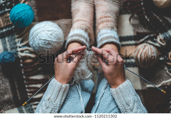 Cozy knitting woman in knitted winter\
warm socks and in pajamas enjoys knit work on brown checkered plaid\
blanket at home in cozy winter time. Top view\
