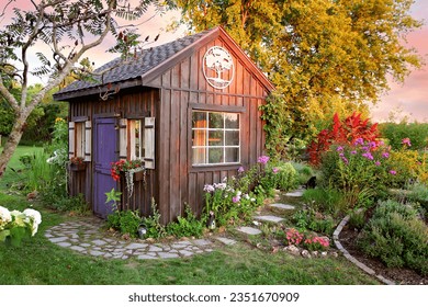 A cozy little wooden cottage garden shed sits in a grassy landscaped bed of flowers at Sunset. - Powered by Shutterstock