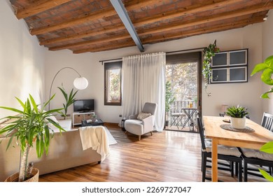 Cozy living area with wood-beamed ceiling with dining table sofas and TV with potted palm tree overlooking the open balcony door. Mediterranean house interior concept
