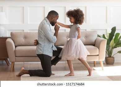 In cozy living room african dad got down on one knee kisses hand of daughter princess wearing fluffy pink skirt, chivalrous gesture, courtesy and politeness, devotion admiration, good manners concept