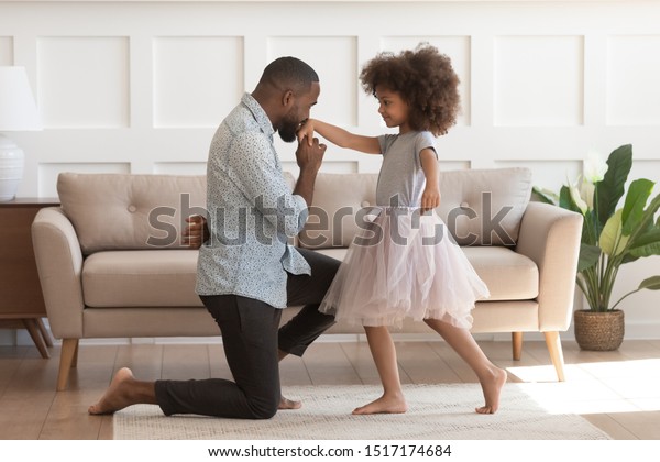 In cozy living room african dad got down on one\
knee kisses hand of daughter princess wearing fluffy pink skirt,\
chivalrous gesture, courtesy and politeness, devotion admiration,\
good manners concept