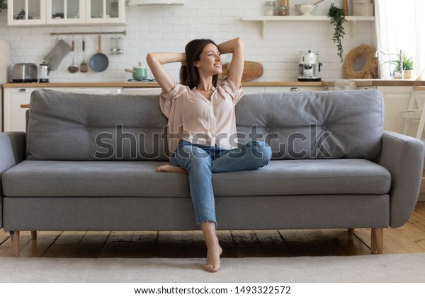 In cozy living room happy woman put hands behind\
head sitting leaned on couch 30s european female enjoy lazy weekend\
or vacation, housewife relaxing feels satisfied accomplish chores\
housework concept