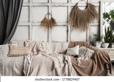 Cozy living room interior in boho chic style with couch, comfortable cushions, bedspread, house decor and green plants in flower pots near mirror on commode