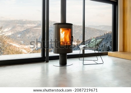 Cozy living room with modern fireplace and panoramic window with great view on the snowy mountains. Concept of rest in houses or cabins on nature. Solitude in nature and escape from everyday life