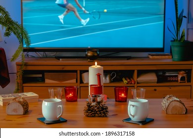 Cozy Living Room Scene With Burning Candles, Coffee Mugs And Decorative Winter Objects With A Hard Court Tennis Match On TV In The Background. Selective Focus On Candles, TV Image Is Blurred.