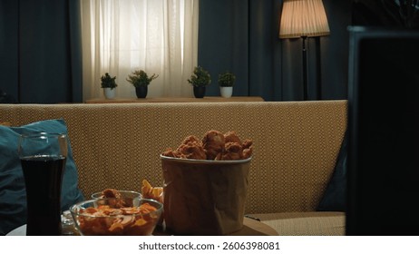 A cozy living room scene with a large bucket of fried chicken, bowls of chips and snacks, and a glass of soda on a table in front of a patterned sofa. Warm lighting adds a relaxed mood.