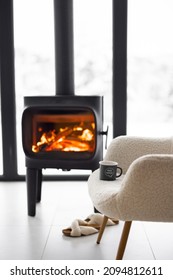 Cozy Living Space By The Burning Fireplace With Chair, Cup And Slippers On Background Of Snowy Landscape. Idea Of Home Comfort And Winter Mood