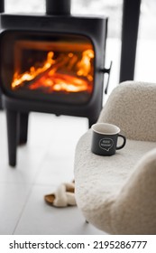 Cozy Living Space By The Burning Fireplace With Chair, Cup And Slippers On Background Of Snowy Landscape. Idea Of Home Comfort And Winter Mood