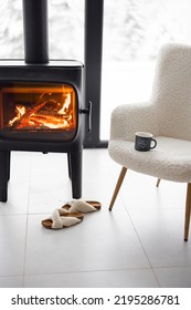 Cozy Living Space By The Burning Fireplace With Chair, Cup And Slippers On Background Of Snowy Landscape. Idea Of Home Comfort And Winter Mood