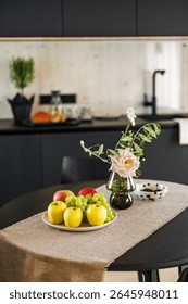 A cozy modern kitchen featuring a table with fresh fruits, a pink rose in a vase, and a dotted ceramic bowl, with a minimalist black and wood interior design