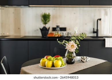 A cozy modern kitchen featuring a table with fresh fruits, a pink rose in a vase, and a dotted ceramic bowl, with a minimalist black and wood interior design