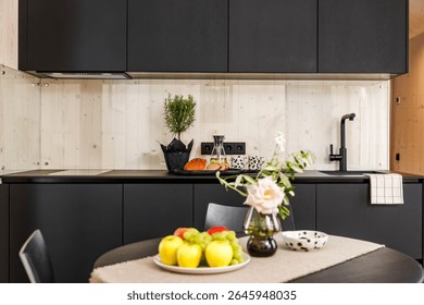 A cozy modern kitchen featuring a table with fresh fruits, a pink rose in a vase, and a dotted ceramic bowl, with a minimalist black and wood interior design