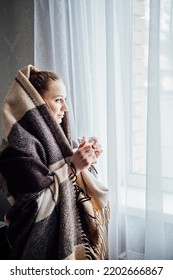 Cozy mood, baby its cold outside. Young woman standing near window with plaid, drinking coffee or tea, looking outside, relaxing in her living room