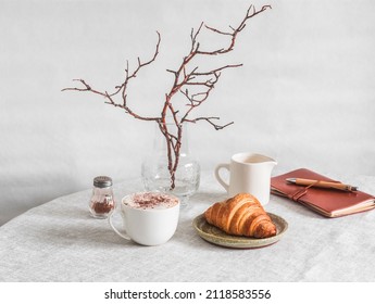 Cozy morning - cappuccino, croissant, vase with branches, leather planner notebook on the table in a bright cozy room. Cozy home interior       