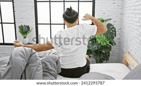 Cozy morning as a handsome young chinese man wakes up, stretches arms back in bed while sitting up, basking in the comfort of his relaxing bedroom in an indoor apartment.