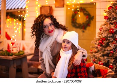 Cozy Outdoor Christmas Photo Of Happy Family Smiling Mother And Little Girl Daughter Sitting Near Festively Decorated House Entrance, Mom With Child Sitting By Xmas Tree Outside. Winter Holidays