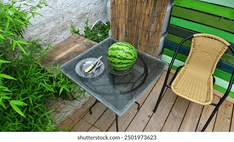 A cozy outdoor patio with a glass table holding a watermelon and cutlery plate set, next to a wicker chair and lush greenery. - Powered by Shutterstock