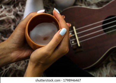 Cozy Photo Of Woman's Hands Holding Cup Of Tea With Ukulele On Backdrop. Fall Or Winter Time Concept. Wellbeing And Simple Pleasures Concept