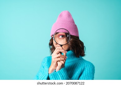 Cozy portrait of a young woman in a knitted blue sweater and a pink hat with bright makeup holding a magnifying glass, fooling around, having fun - Powered by Shutterstock