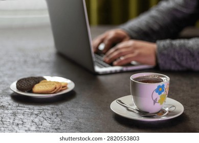 Cozy remote work setup featuring hot chocolate mug, biscuits, and a person typing on a laptop in soft focus