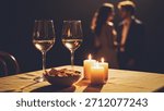 A cozy romantic dinner setup with two wine glasses, candles, and a couple in the background.