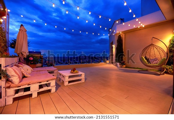 cozy rooftop patio area\
with lounge zone, hanging chair and and string lights at warm\
summer evening