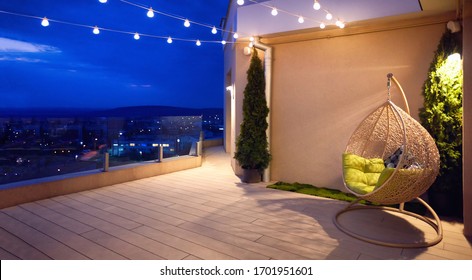 gemütliche Dachterrasse mit Rattanhängestuhl, Garland und schöner Landschaft am Abend