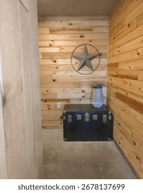 A cozy, rustic interior corner featuring a black vintage trunk with silver hardware, a modern metal vase with a green plant, and a decorative metal star mounted on natural wood paneling. 