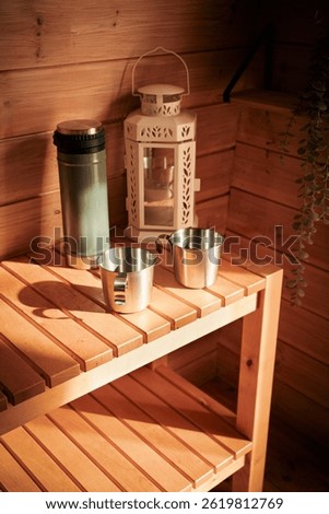 Cozy rustic scene featuring two stainless steel mugs, a green thermos, and a decorative white lantern placed on a wooden shelf. 