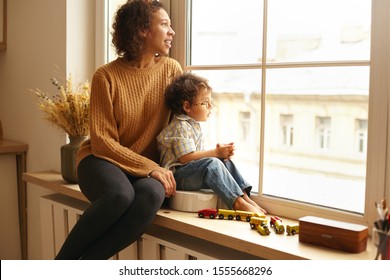 Cozy Scene Of Joyful Young Latin Female Wearing Jeans And Knitted Jumper Sitting On Windowsill Hugging Her Cute Chubby Little Child, Both Watching Outside, Talking, Having Dreamy Thoughtful Expression