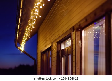 Cozy Scene Of String Lights Hanging Under The Roof At Evening Time. Fashion Decoration With Bulbs For Night Summer Or Christmas Party. Outdoor Electric Lamps