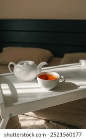 A cozy setup with a cup of hot tea and a teapot on a white tray placed on a bed.
