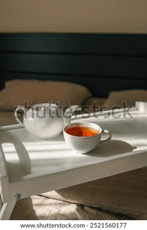 A cozy setup with a cup of hot tea and a teapot on a white tray placed on a bed.