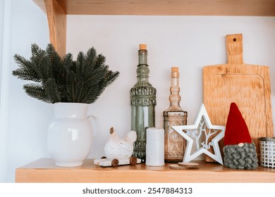 A cozy shelf displays a white pitcher with green foliage, vintage glass bottles, and festive decorations, creating a warm, inviting atmosphere perfect for the holiday season. - Powered by Shutterstock