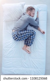 Cozy Sleep. Top View On A Young Man Wearing Pajamas Embracing His Pillow While Sleeping Tight In A Fetal Position At Home.