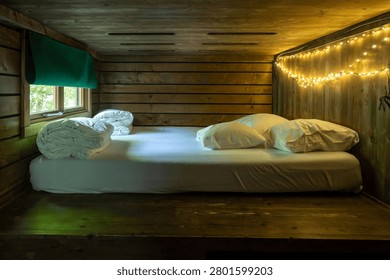 Cozy sleeping loft in a rustic forest cabin with white bedding, plump pillows and warm fairy lights strung along the wooden wall. A small window framed by a green blind reveals the forest, creating an intimate off-grid nature escape for restful nights